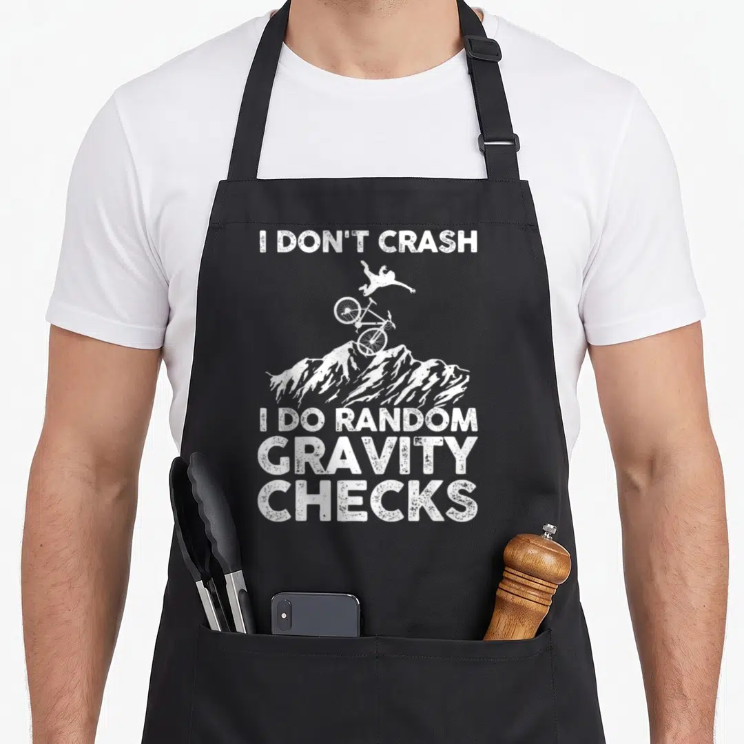 Mtb biking bike apron - White distressed text I Don't Crash Random Gravity Checks with airborne biker over mountain peak — shown on a black cotton canvas apron against a white background