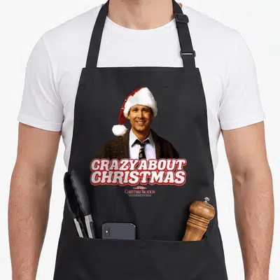 Funny national lampoon's christmas vacation apron - Smiling man in Santa hat above red and white CRAZY ABOUT CHRISTMAS — shown on a black cotton canvas apron against a white background