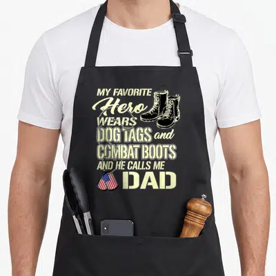 Dog tag apron - Pale yellow text reading My Favorite Hero Wears Dog Tags and Combat Boots And He Calls Me Dad — shown on a black cotton canvas apron against a white background