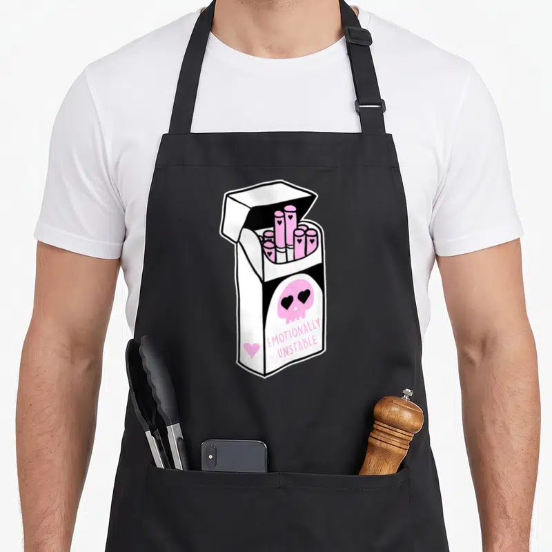 Emotionally goth apron - Open cigarette pack with pink cigarettes, pink skull with heart eyes, and EMOTIONALLY UNSTABLE — shown on a black cotton canvas apron against a white background