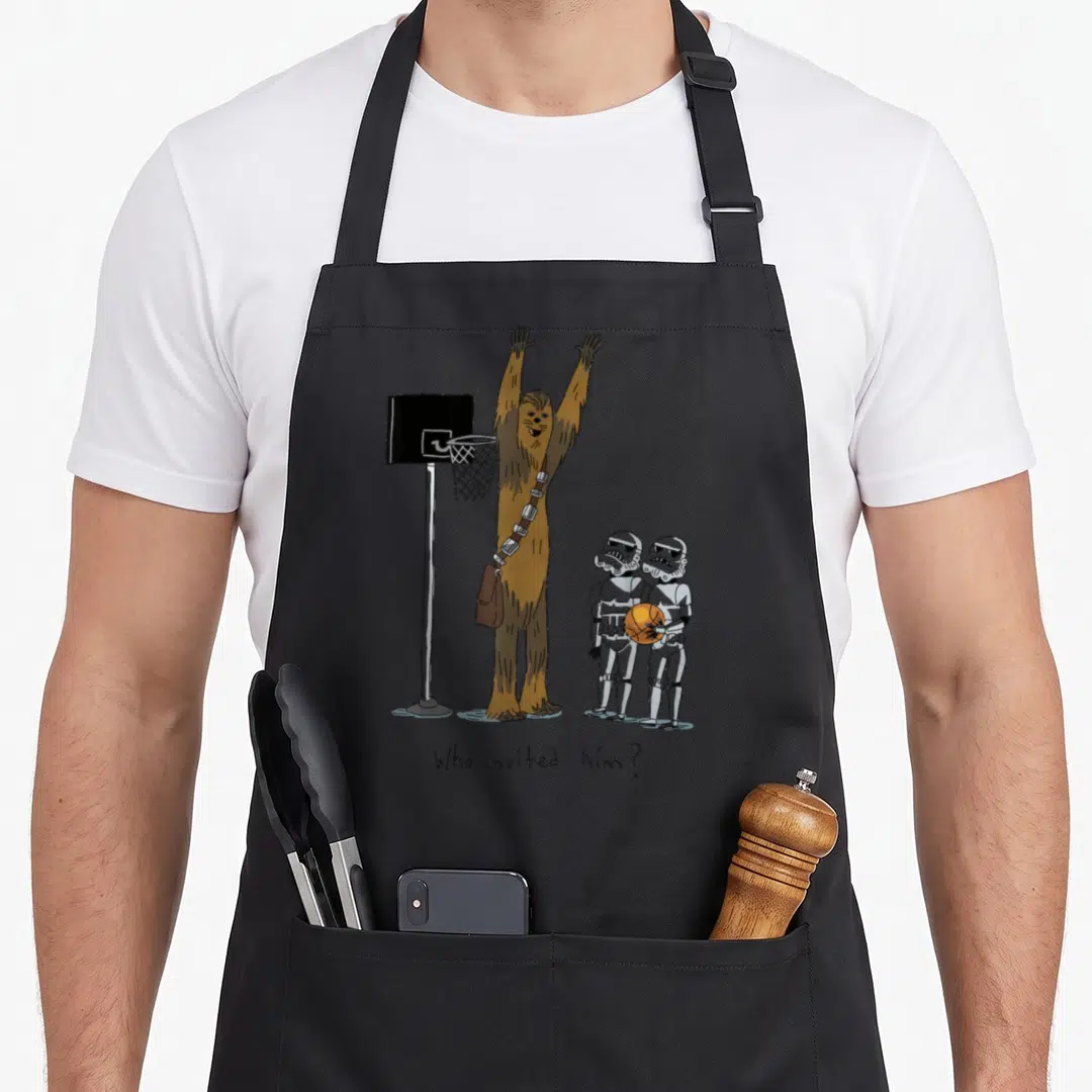 Basketball crazy falcon man apron - Chewbacca raising arms by hoop, two Stormtroopers holding orange ball, text below — shown on a black cotton canvas apron against a white background