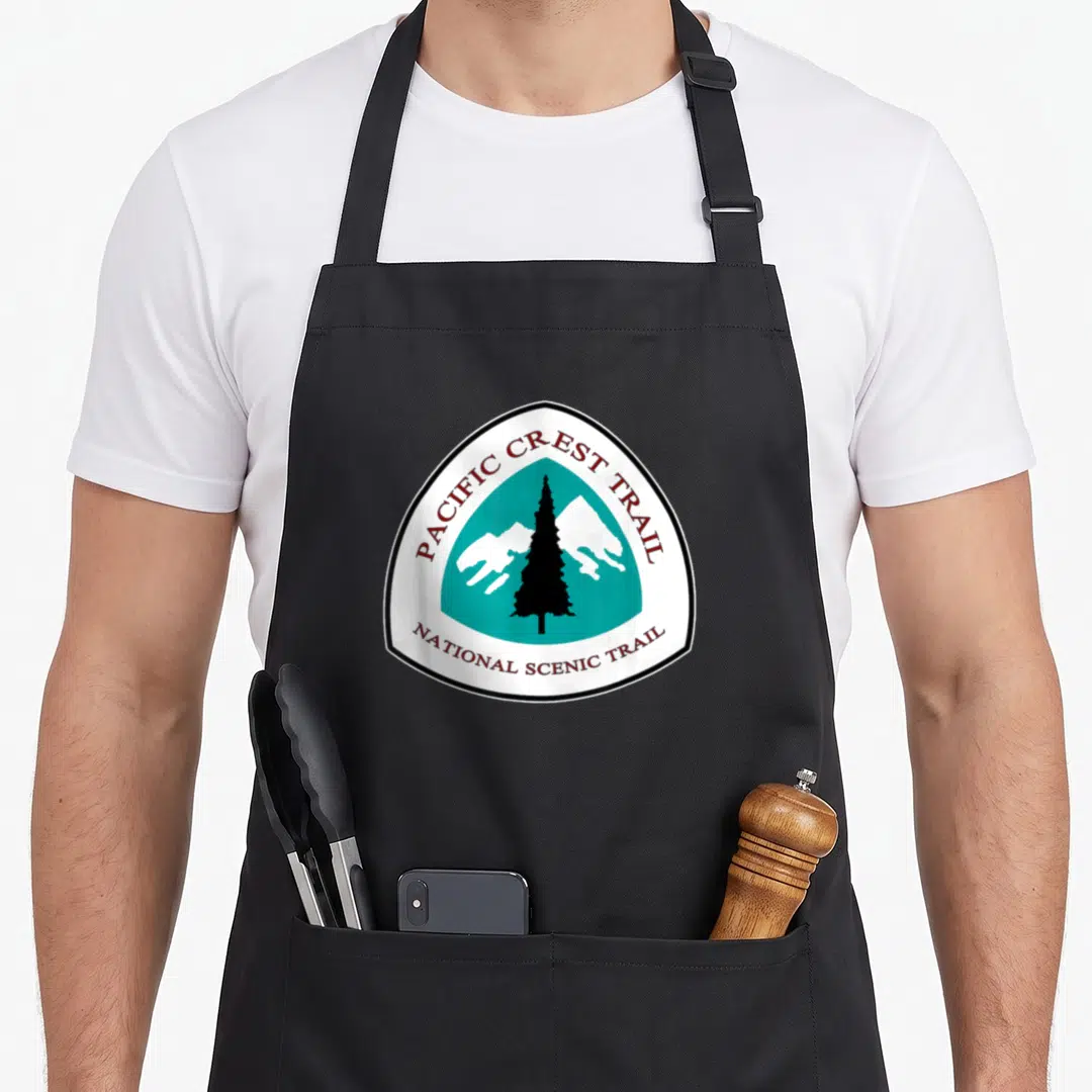 Rangertee backpacking apron - Shield logo with black pine tree, white mountains, and PCT text on teal background — shown on a black cotton canvas apron against a white background