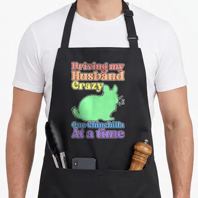 Chinchilla driving apron - Green chinchilla silhouette with rainbow stacked text reading Driving my Husband Crazy One — shown on a black cotton canvas apron against a white background