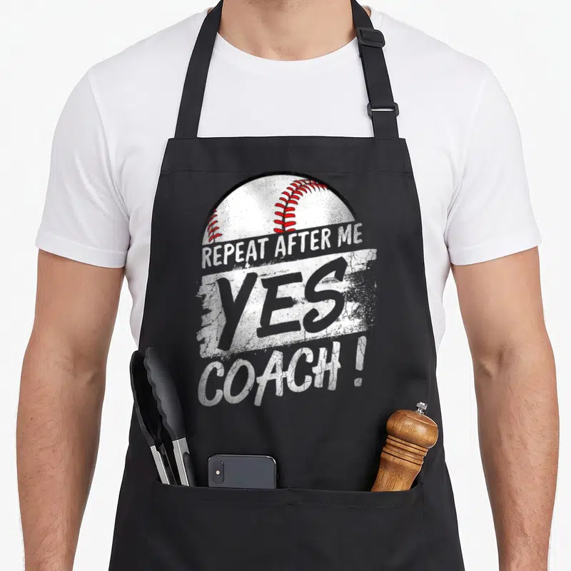 After baseball apron - Distressed white and red text reading REPEAT AFTER ME YES COACH! over a baseball graphic on grey — shown on a black cotton canvas apron against a white background