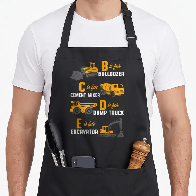 Construction heavy canvas aprons - B is for BULLDOZER, C is for CEMENT MIXER, D is for DUMP TRUCK, E is for EXCAVATOR — shown on a black cotton canvas apron against a white background
