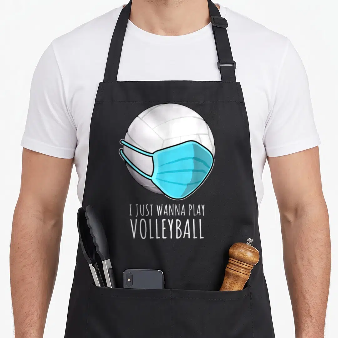 Play volleyball apron - Cartoon volleyball wearing a light blue face mask above white text reading I JUST WANNA PLAY — shown on a black cotton canvas apron against a white background