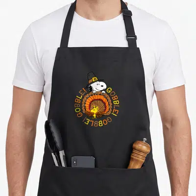 Woodstock gobble thanksgiving apron - Snoopy in pilgrim hat peeking over woodstock in turkey feathers with circular — shown on a black cotton canvas apron against a white background