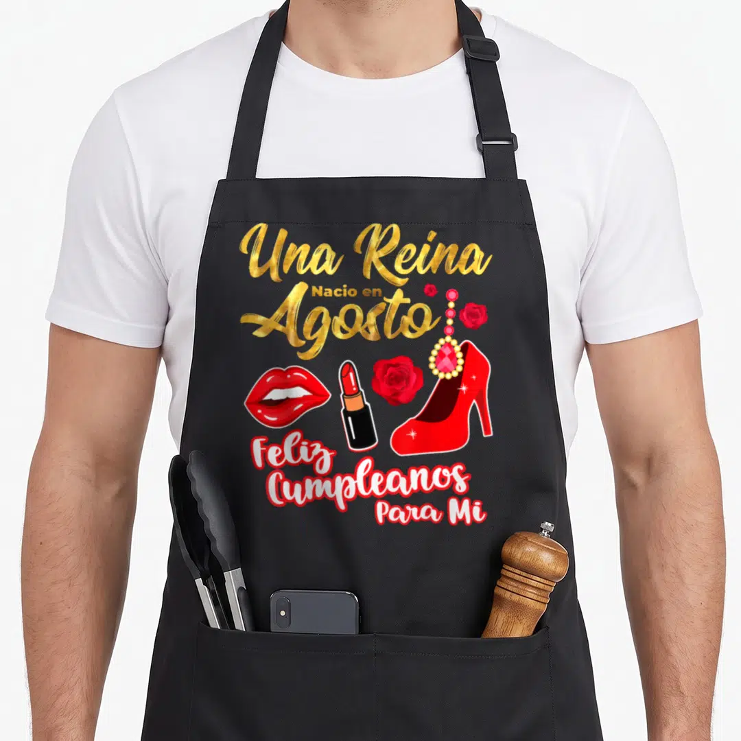 Una birthday apron - Gold Una Reina Nacio en Agosto text above red high heel, lipstick, lips, rose, and earring cluster — shown on a black cotton canvas apron against a white background