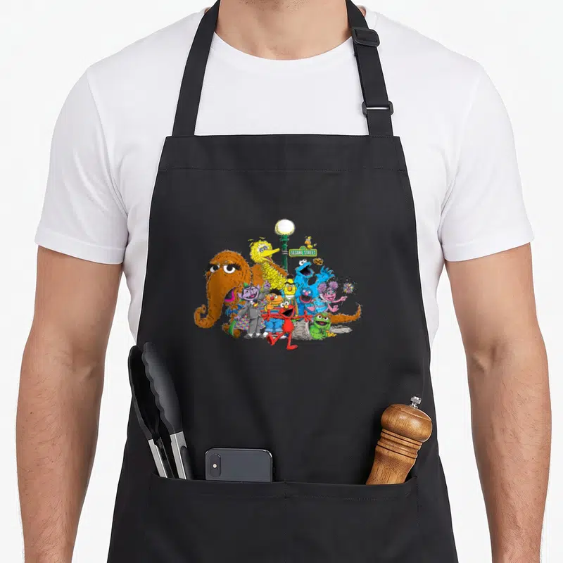 Light street apron - Sesame Street characters clustered around a green lamp post with sign in orange yellow blue red — shown on a black cotton canvas apron against a white background