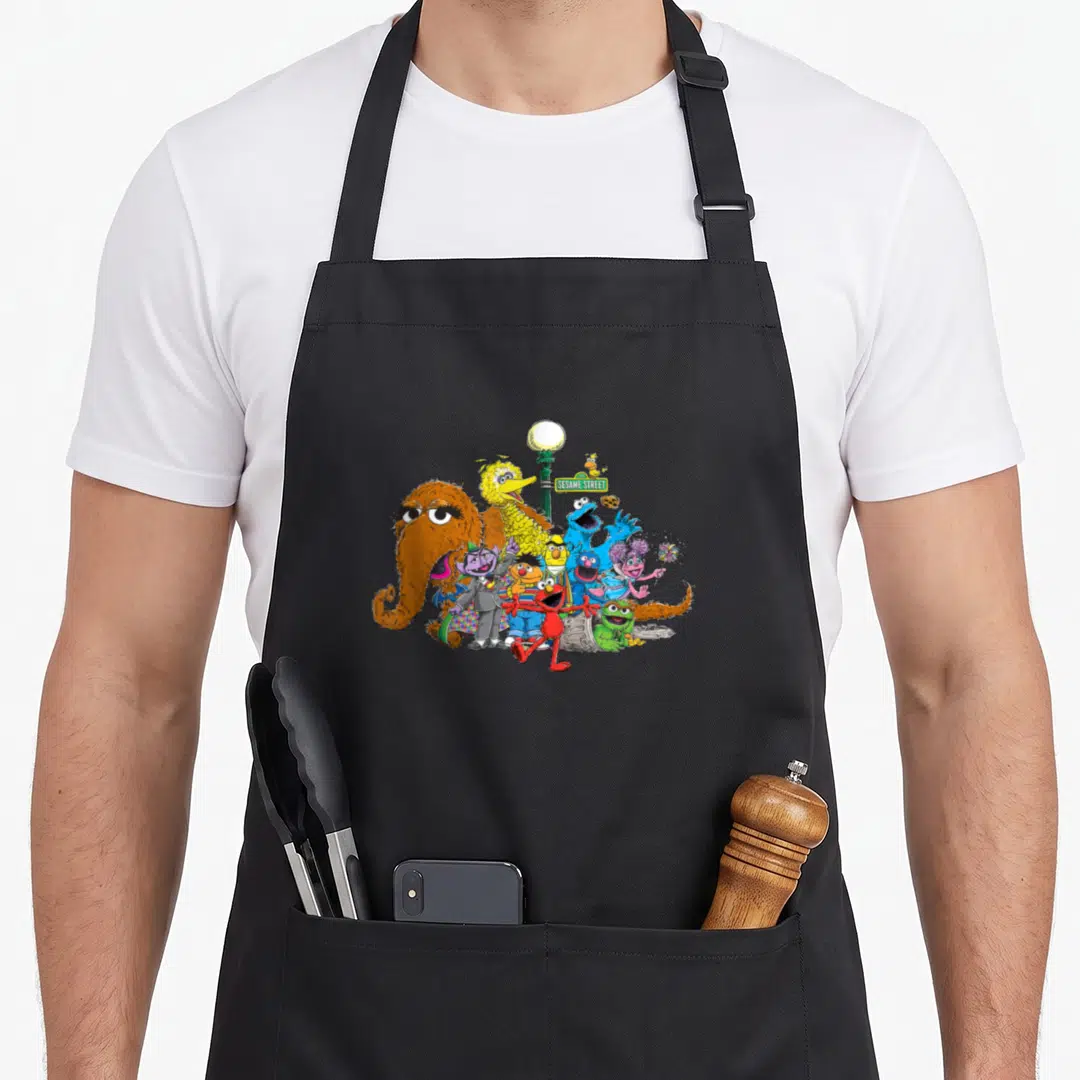 Light street apron - Sesame Street characters clustered around a green lamp post with sign in orange yellow blue red — shown on a black cotton canvas apron against a white background