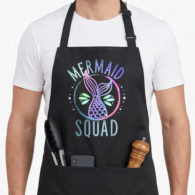 Mermaid squad family aprons - Watercolor MERMAID SQUAD text with pink purple blue tail and green leaves in a circle — shown on a black cotton canvas apron against a white background