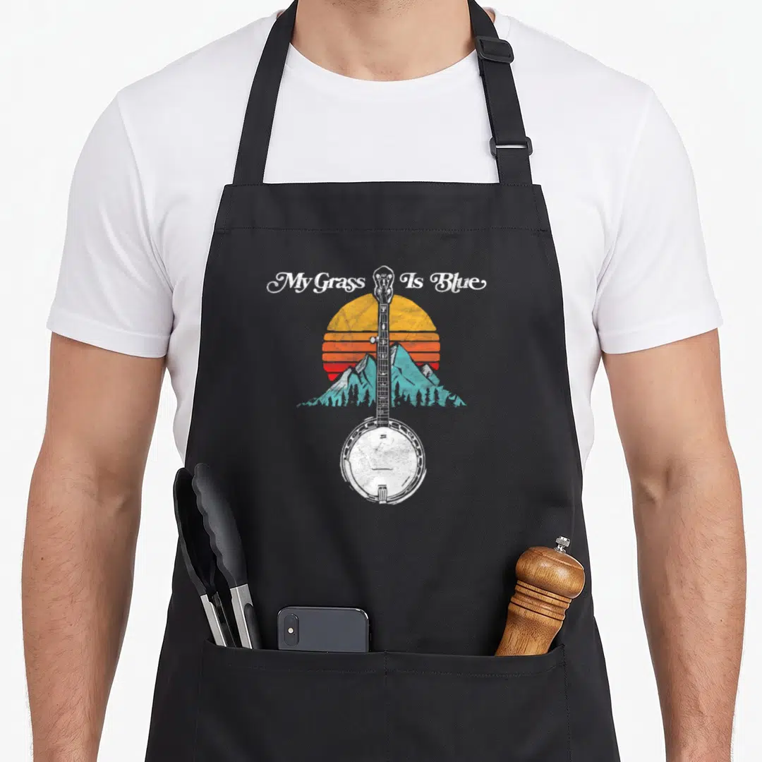 Grass bluegrass apron - Retro banjo illustration with My Grass Is Blue text over striped sun and teal mountains — shown on a black cotton canvas apron against a white background