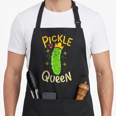 Pickle vegetable apron - Cartoon green pickle wearing a gold crown surrounded by hearts and sparkles — shown on a black cotton canvas apron against a white background