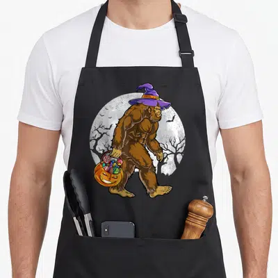 Bigfoot halloween kitchen aprons - Brown bigfoot in purple witch hat carrying orange pumpkin candy bucket near white — shown on a black cotton canvas apron against a white background