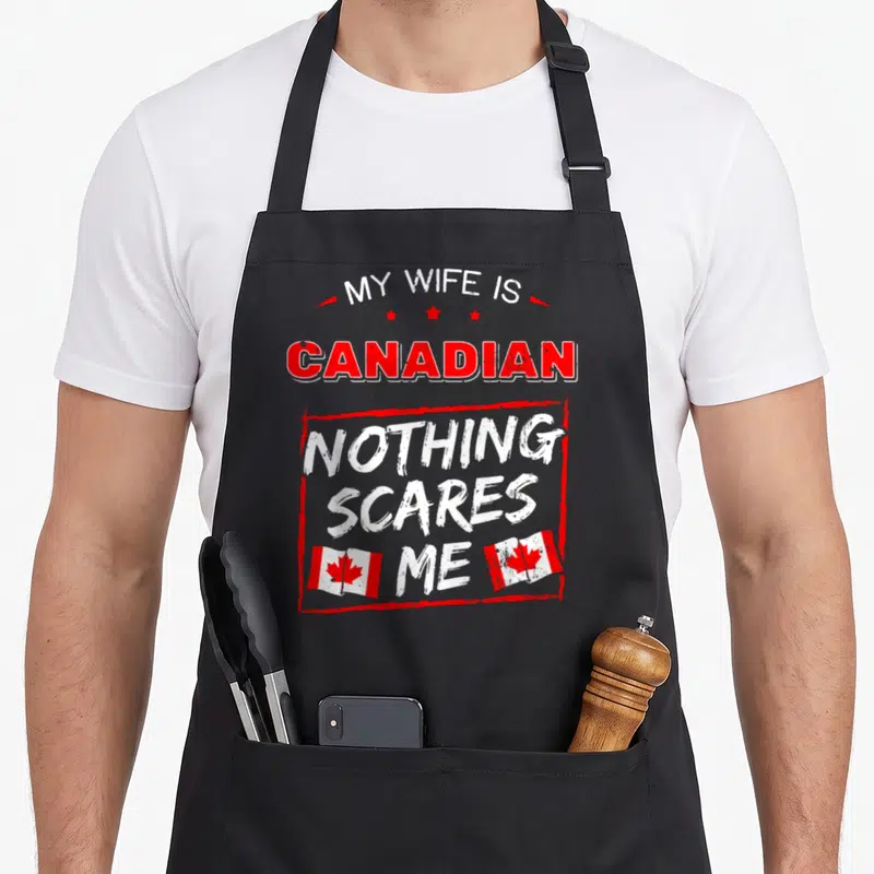 Canadian wife apron - Red and white stacked text reading My Wife Is Canadian Nothing Scares Me with three stars and two — shown on a black cotton canvas apron against a white background