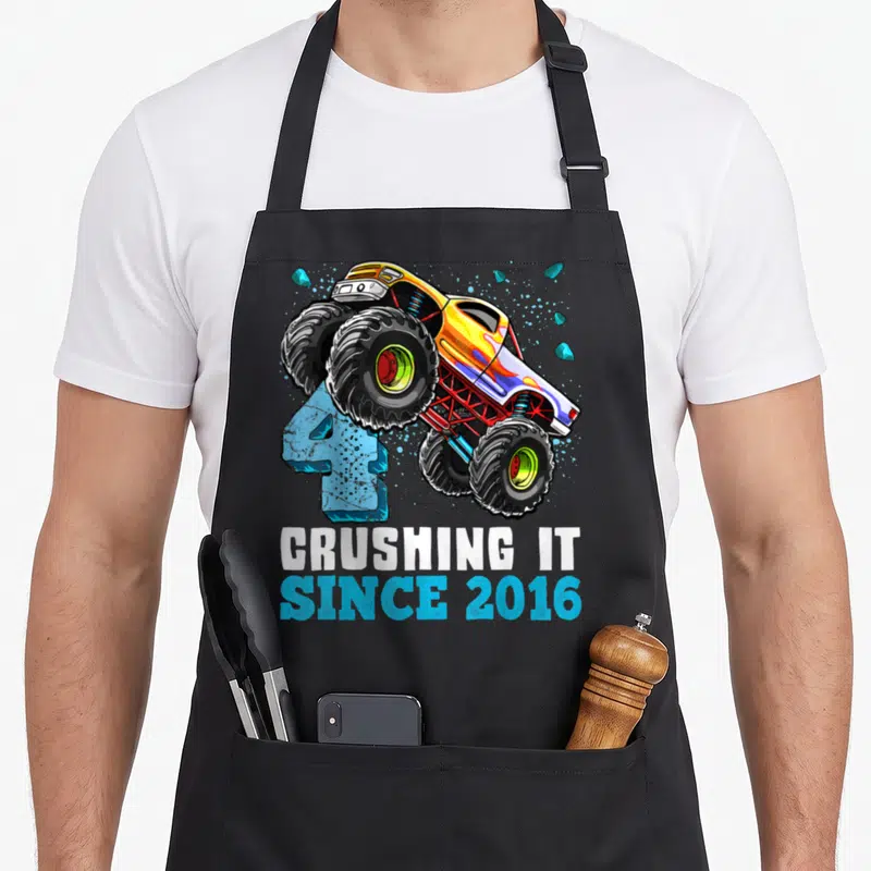 Crushing monster 4th truck apron - Yellow monster truck jumping over blue number 4 with stacked white and blue text — shown on a black cotton canvas apron against a white background