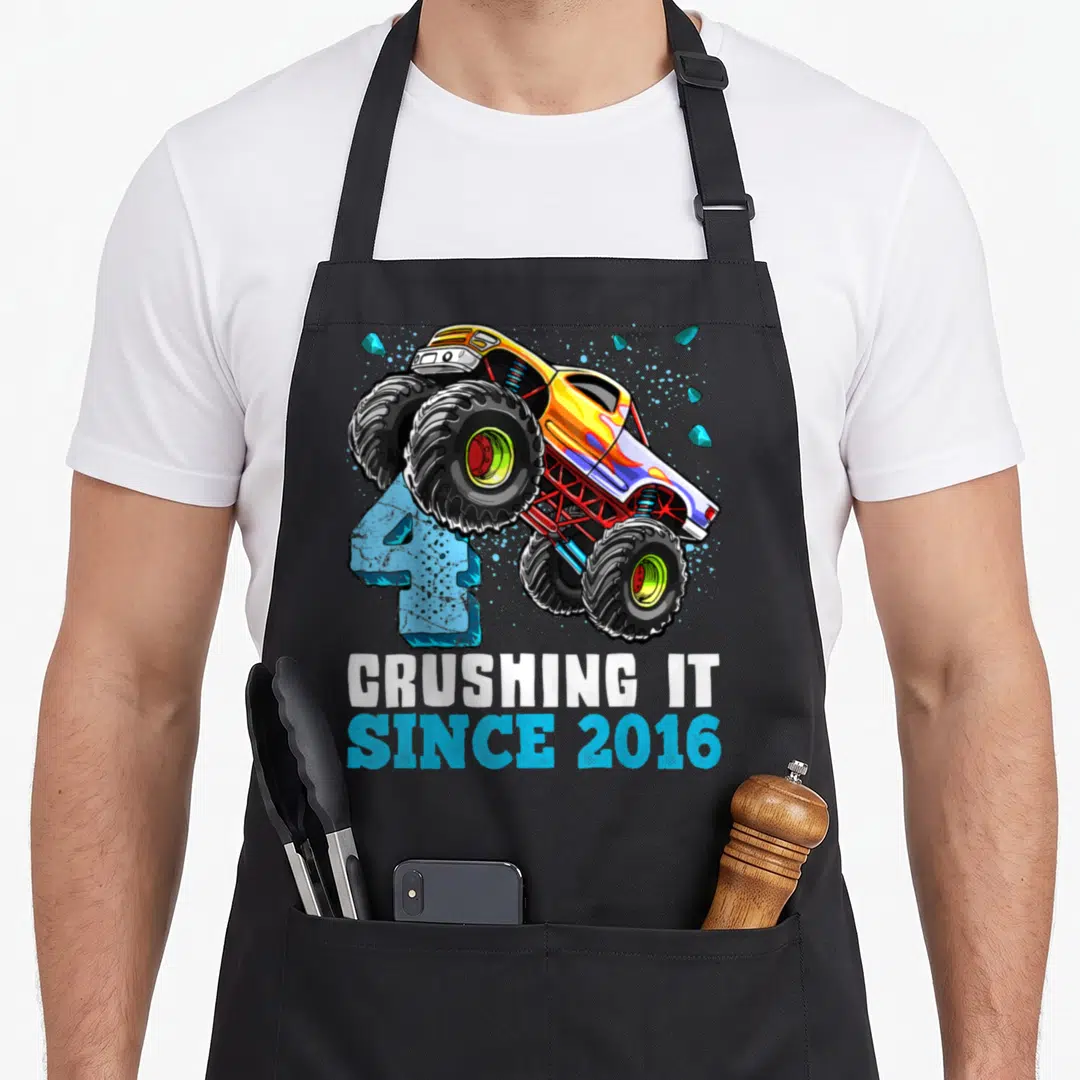 Crushing monster 4th truck apron - Yellow monster truck jumping over blue number 4 with stacked white and blue text — shown on a black cotton canvas apron against a white background