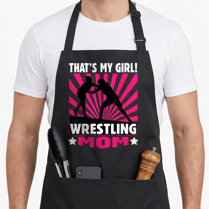 Cute wrestling apron - That's My Girl text, two wrestler silhouettes, pink sunburst, Wrestling Mom label — shown on a black cotton canvas apron against a white background
