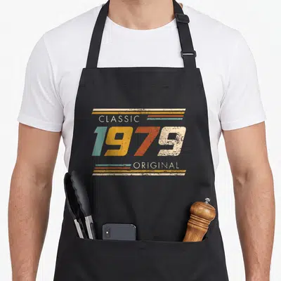 Original est birthday apron - Retro 1979 text in teal, yellow, orange, and beige with horizontal stripes and distressed — shown on a black cotton canvas apron against a white background