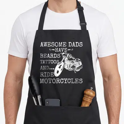 Motorcycle tattoo aprons - White distressed text listing beards tattoos and motorcycles over a bike graphic on black — shown on a black cotton canvas apron against a white background