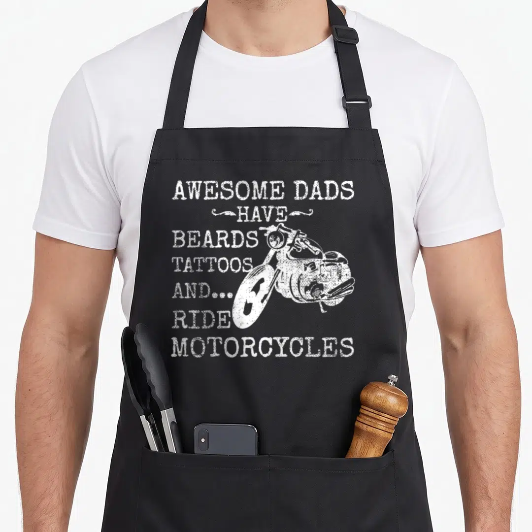 Motorcycle tattoo aprons - White distressed text listing beards tattoos and motorcycles over a bike graphic on black — shown on a black cotton canvas apron against a white background