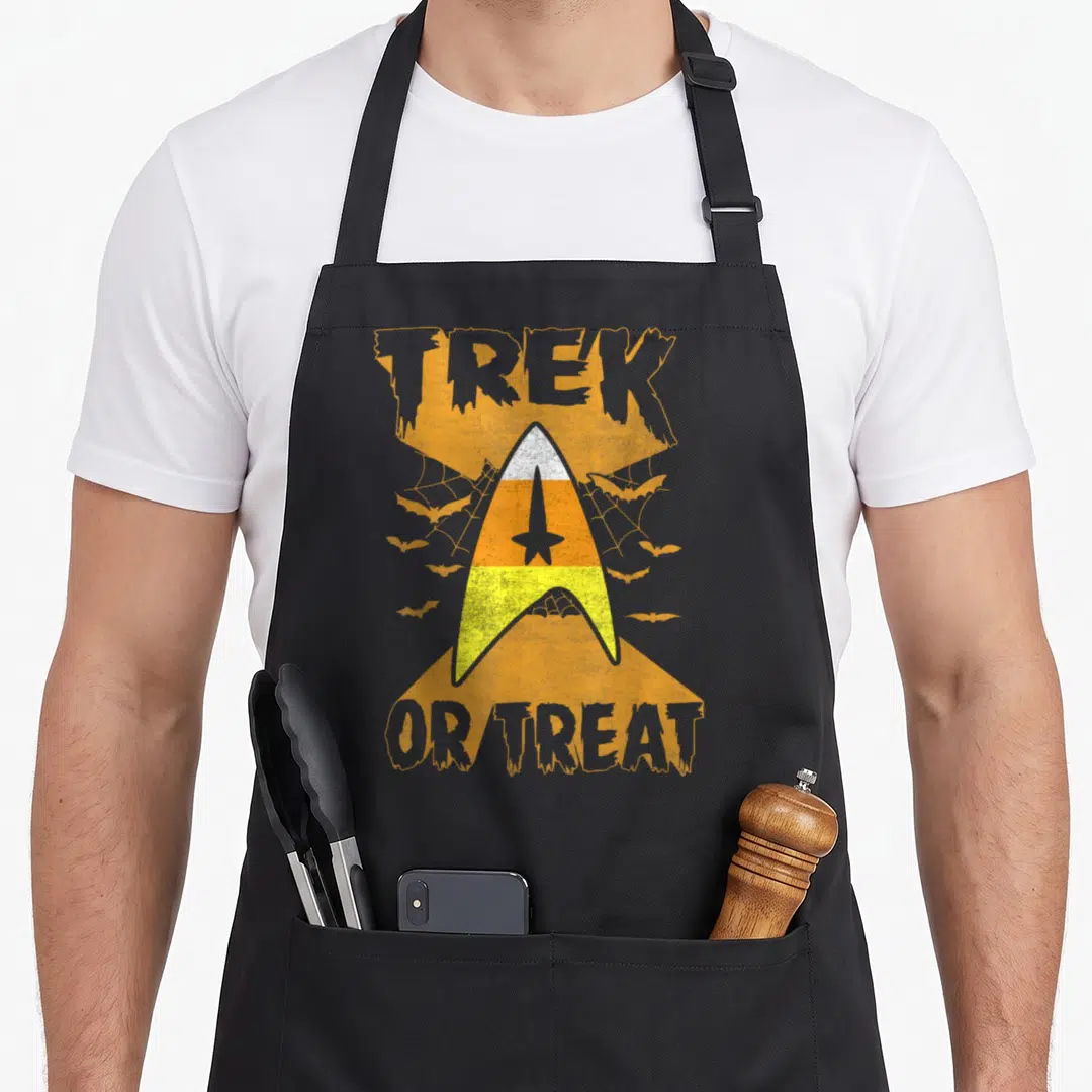 Corn treat halloween apron - Distressed Starfleet insignia in candy corn colors with TREK OR TREAT text and bats — shown on a black cotton canvas apron against a white background