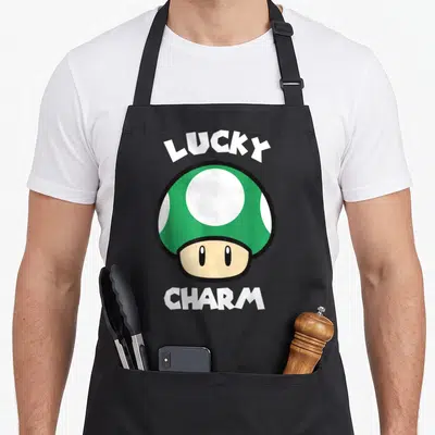 Patty mushroom apron - Cartoon mushroom with LUCKY CHARM text in green and white — shown on a black cotton canvas apron against a white background