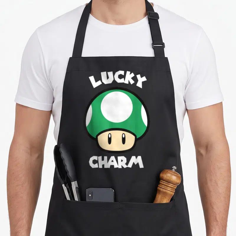 Patty mushroom apron - Cartoon mushroom with LUCKY CHARM text in green and white — shown on a black cotton canvas apron against a white background
