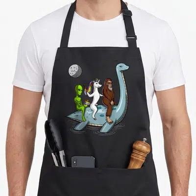 Cryptozoology bigfoot apron - Blue plesiosaur carries green alien with rainbow flag, white unicorn, and brown Bigfoot — shown on a black cotton canvas apron against a white background