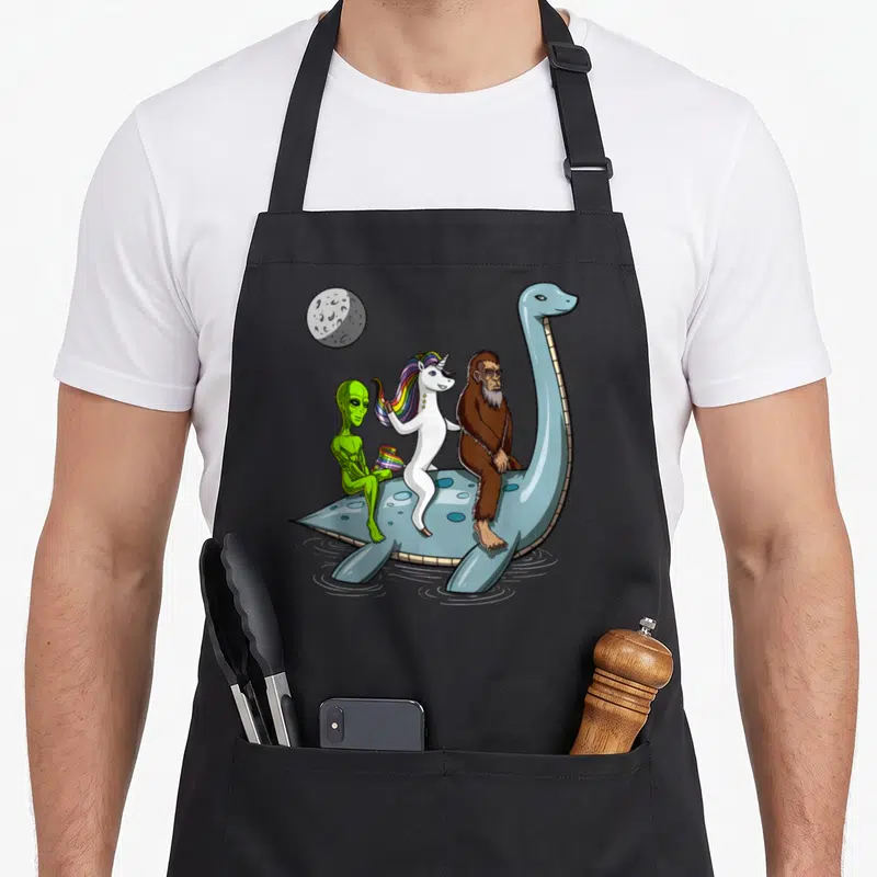 Cryptozoology bigfoot apron - Blue plesiosaur carries green alien with rainbow flag, white unicorn, and brown Bigfoot — shown on a black cotton canvas apron against a white background
