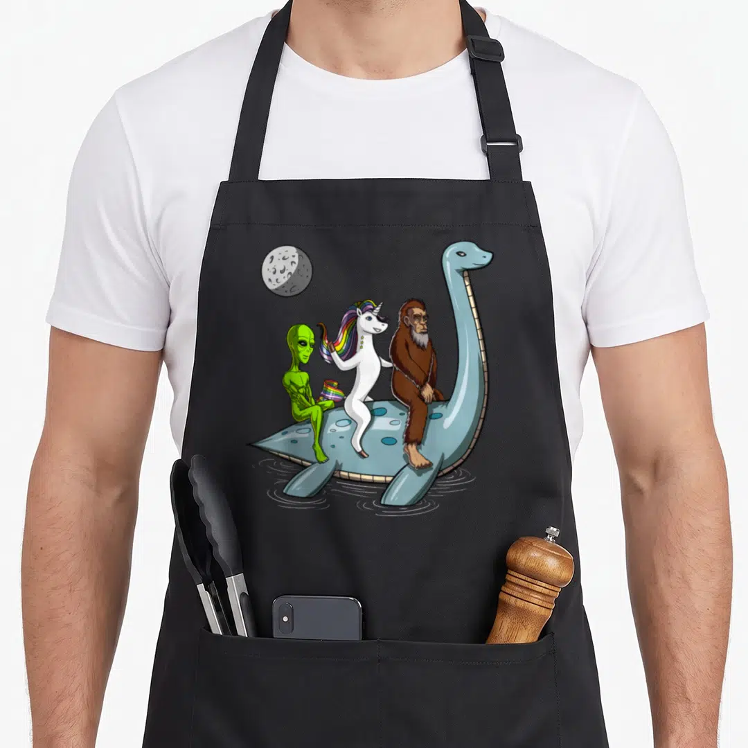 Cryptozoology bigfoot apron - Blue plesiosaur carries green alien with rainbow flag, white unicorn, and brown Bigfoot — shown on a black cotton canvas apron against a white background
