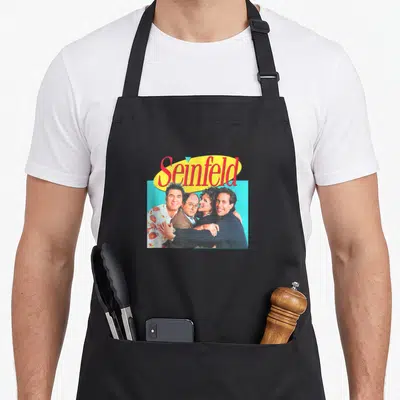 Hugging x apron - Seinfeld logo above photo of four cast members clustered on blue background — shown on a black cotton canvas apron against a white background