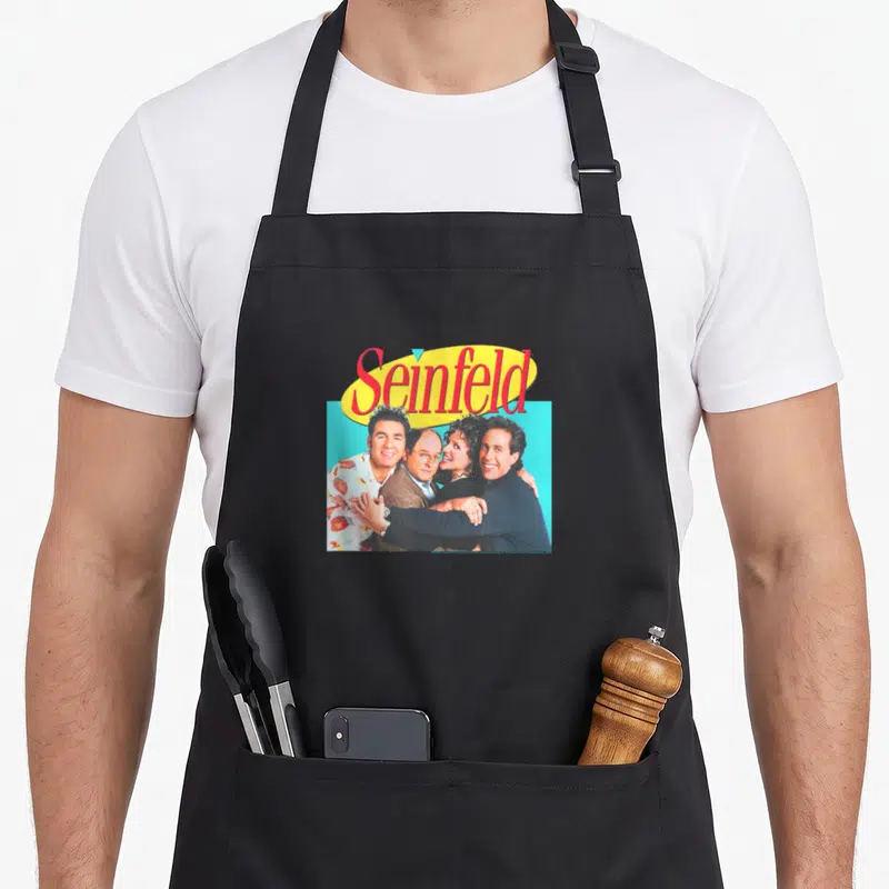 Hugging x apron - Seinfeld logo above photo of four cast members clustered on blue background — shown on a black cotton canvas apron against a white background