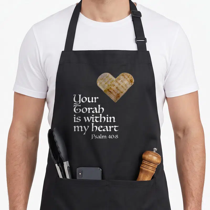 Torah bible verse apron - Heart-shaped stone with Hebrew text above stacked white English lettering reading Your Torah — shown on a black cotton canvas apron against a white background