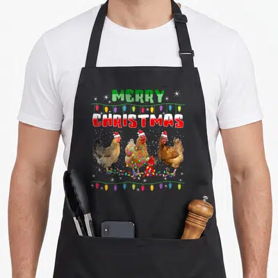 Chicken merry christmas apron - Three chickens in santa hats under merry christmas text with string lights and red — shown on a black cotton canvas apron against a white background