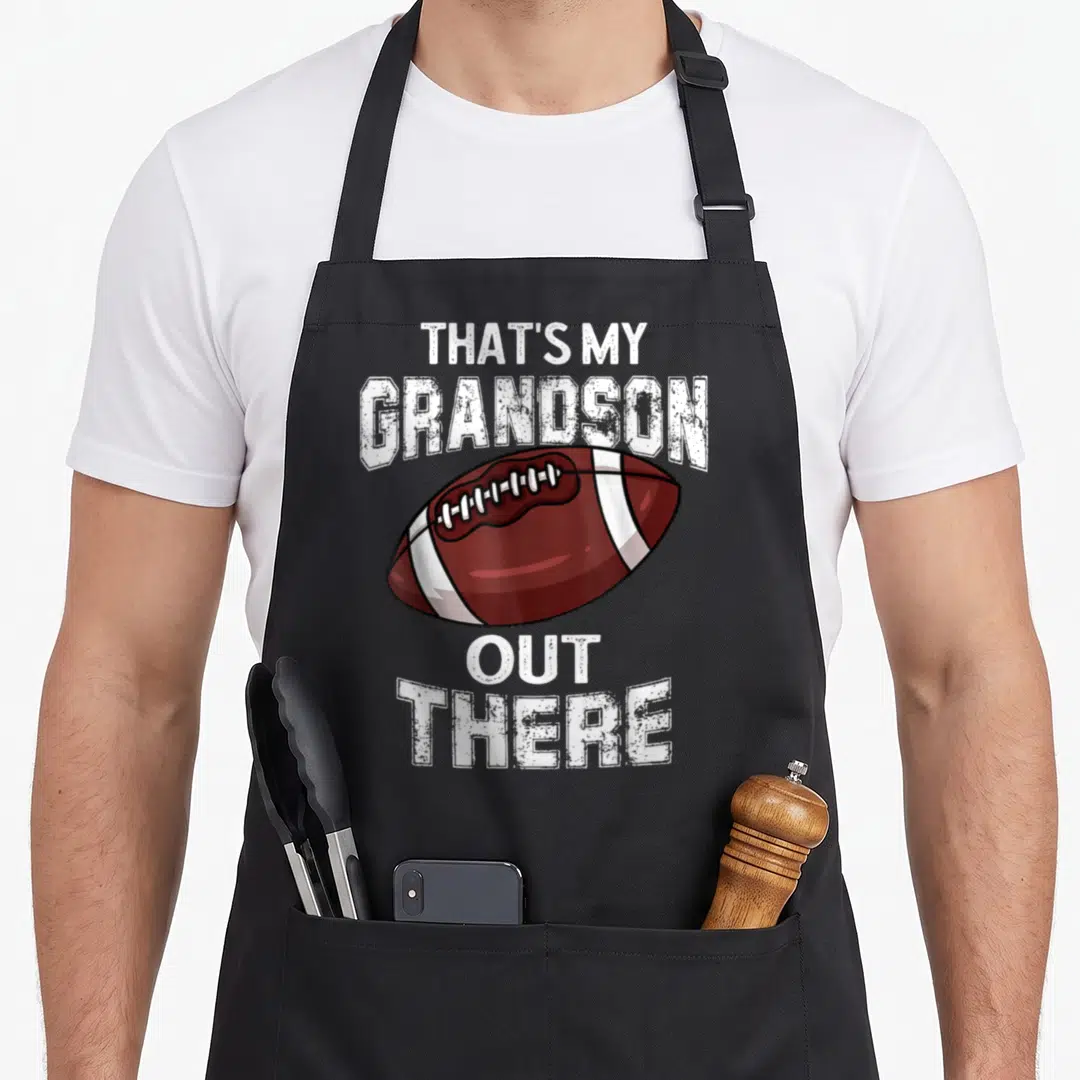 Grandpa grandma aprons - Brown football with white stripes and distressed white text reading That's My Grandson Out — shown on a black cotton canvas apron against a white background