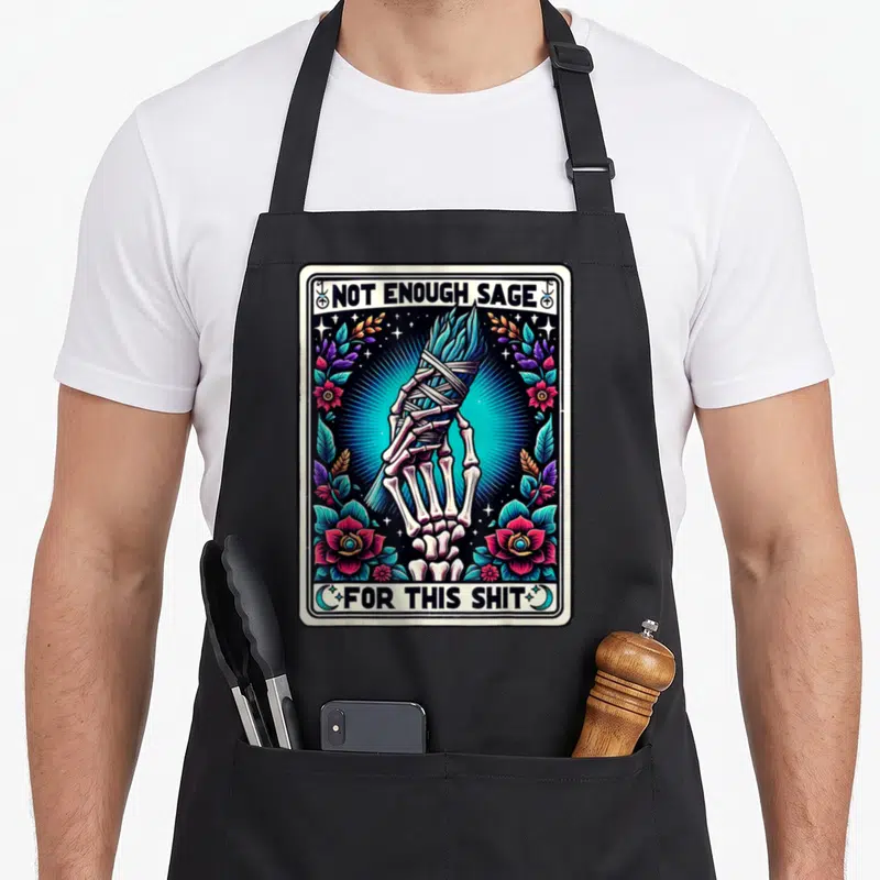 Tarot sage apron - Skeletal hand holding burning bundle surrounded by flowers and stars with text NOT ENOUGH SAGE — shown on a black cotton canvas apron against a white background