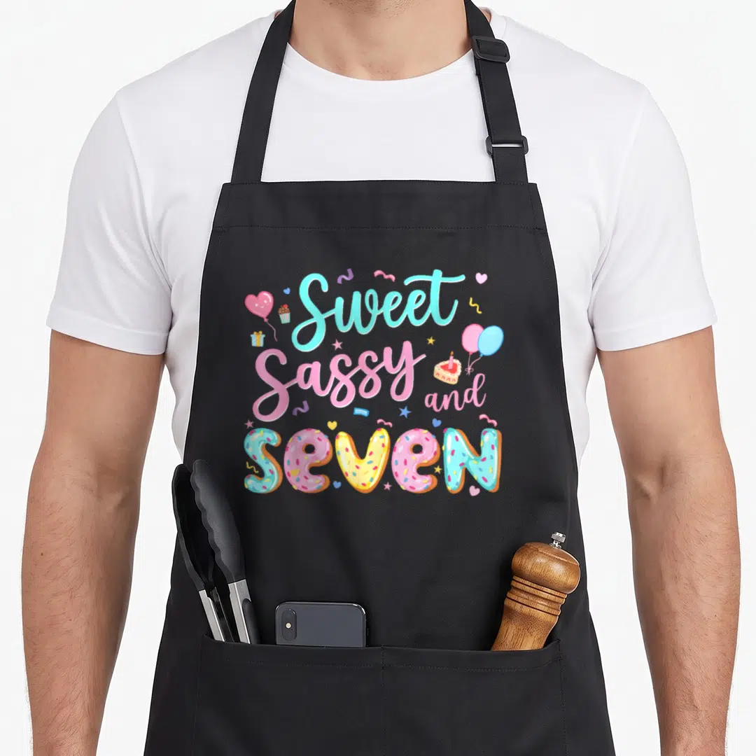 7th birthday sassy apron - Sweet Sassy and Seven donut text with pink blue yellow balloons and cake — shown on a black cotton canvas apron against a white background