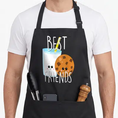 Bestie food aprons - Cartoon milk glass and smiling cookie with BEST FRIENDS text in white, brown, yellow, and blue — shown on a black cotton canvas apron against a white background