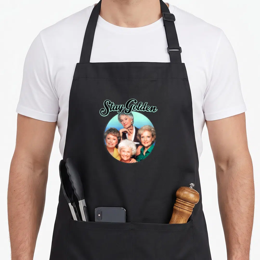 Family golden apron - Stay Golden text arched over circular photo of four women in colorful tops — shown on a black cotton canvas apron against a white background