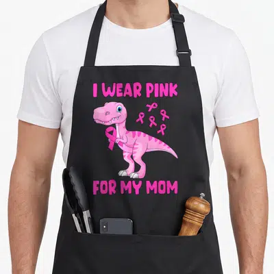 Toddler son breast cancer awareness aprons - Pink cartoon dinosaur holding ribbon with I WEAR PINK FOR MY MOM text — shown on a black cotton canvas apron against a white background
