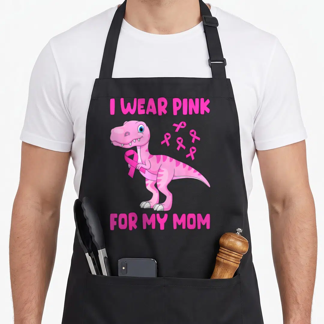 Toddler son breast cancer awareness aprons - Pink cartoon dinosaur holding ribbon with I WEAR PINK FOR MY MOM text — shown on a black cotton canvas apron against a white background