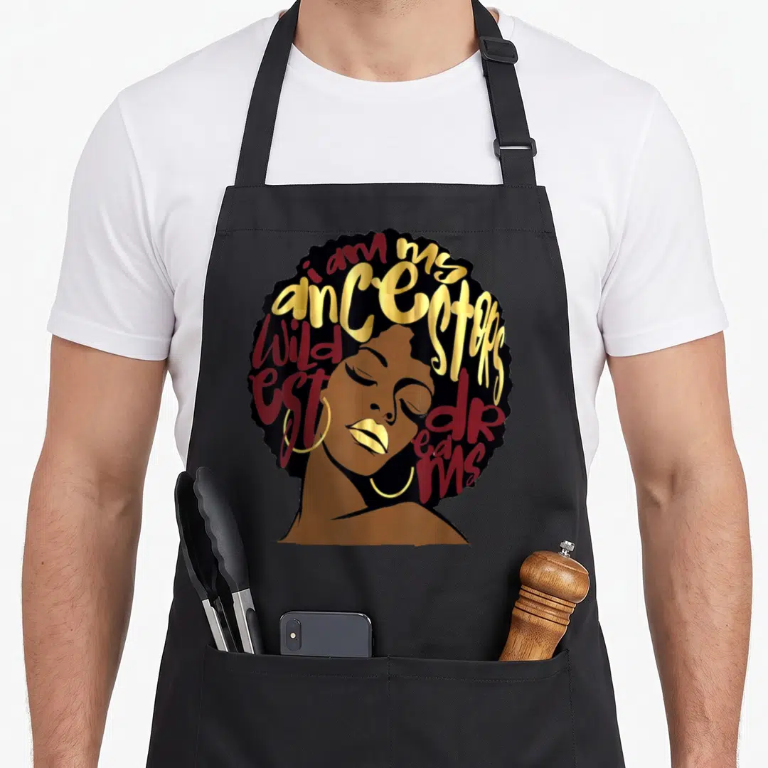 Ancestor the ruby apron - Cartoon woman with brown skin and gold lips; text i am my ancestors wildest dreams forms — shown on a black cotton canvas apron against a white background
