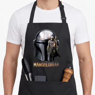 Helmet ivorian apron - Mandalorian standing figure before large helmet with THE MANDALORIAN title text in gold and grey — shown on a black cotton canvas apron against a white background