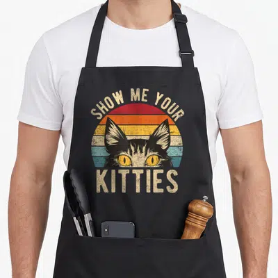 Raunchy kitty cat apron - SHOW ME YOUR KITTIES text over striped sun with black cat head peeking through retro colors — shown on a black cotton canvas apron against a white background