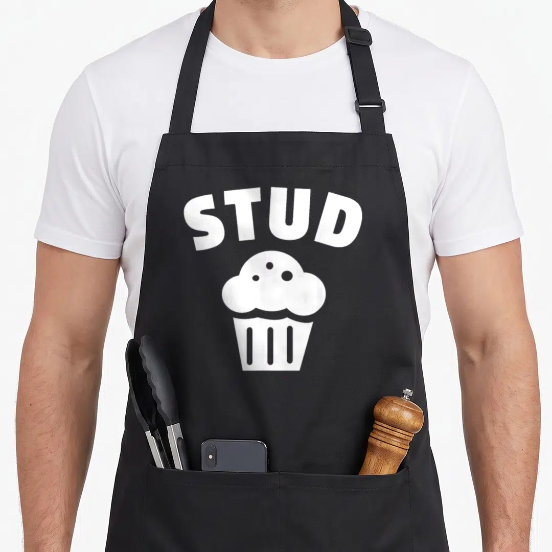 Vintage stud muffin man apron - White arched STUD text above a minimalist cupcake illustration — shown on a black cotton canvas apron against a white background