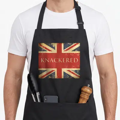 Tired flag funny apron - Distressed Union Jack with KNACKERED text on red band — shown on a black cotton canvas apron against a white background