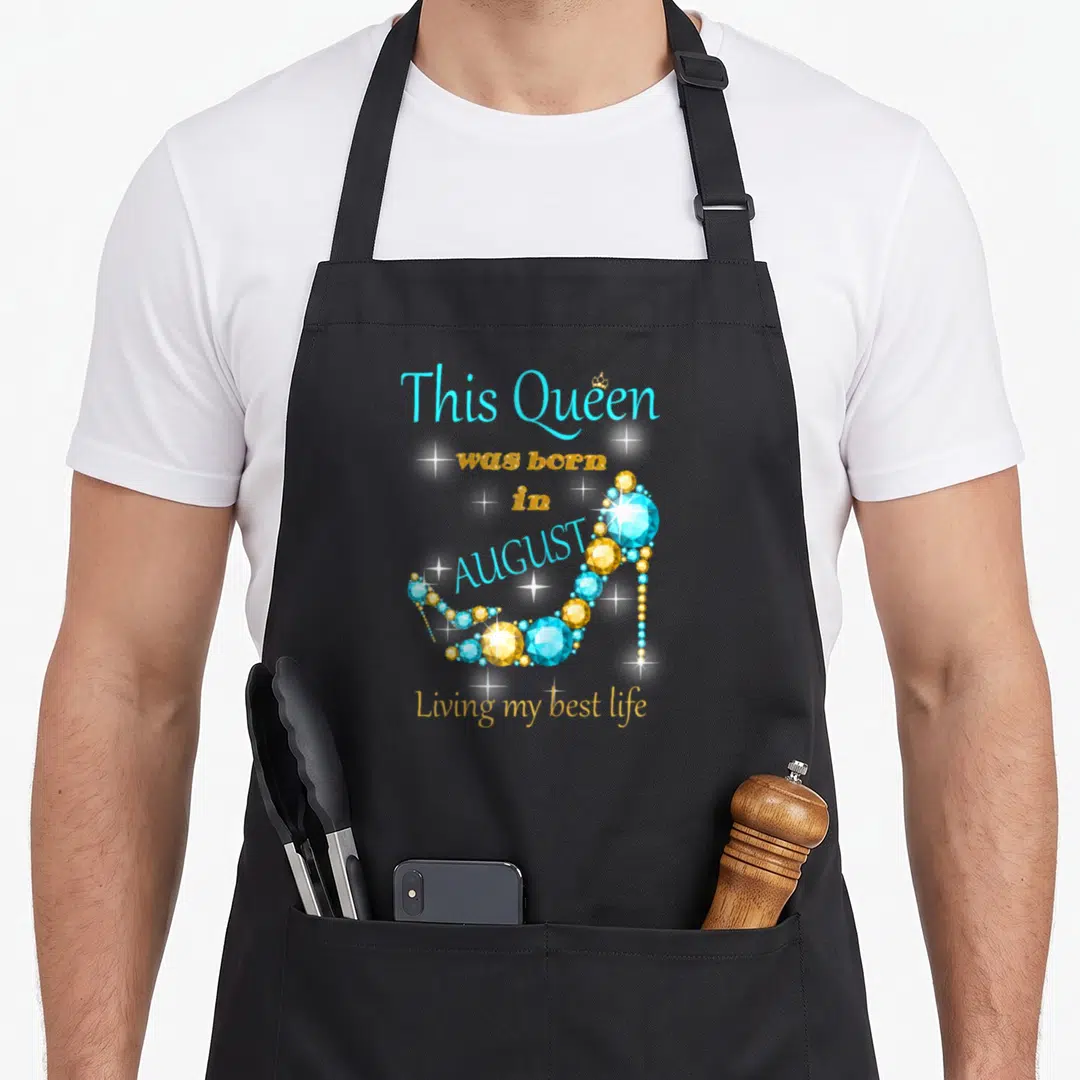 August baking queen apron - Gem high heel shoe with text This Queen was born in August Living my best life — shown on a black cotton canvas apron against a white background