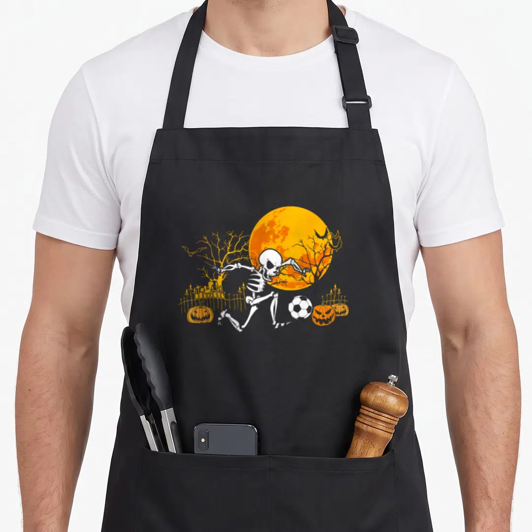 Skeleton player halloween apron - White skeleton kicking soccer ball under orange moon with pumpkins and bats — shown on a black cotton canvas apron against a white background