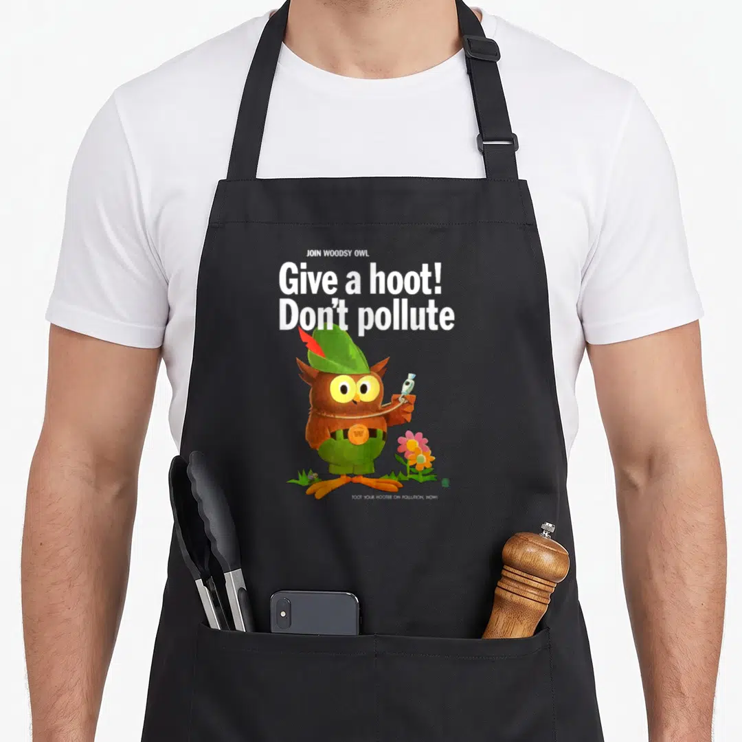 70s owl apron - Cartoon owl holding trash with Give a hoot Don't pollute text in brown and green — shown on a black cotton canvas apron against a white background